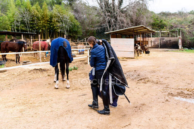 Chica adolescente de cuerpo entero caminando en el paddock y llevando manta de caballo mientras cuida de los animales después de entrenar en la escuela ecuestre - foto de stock