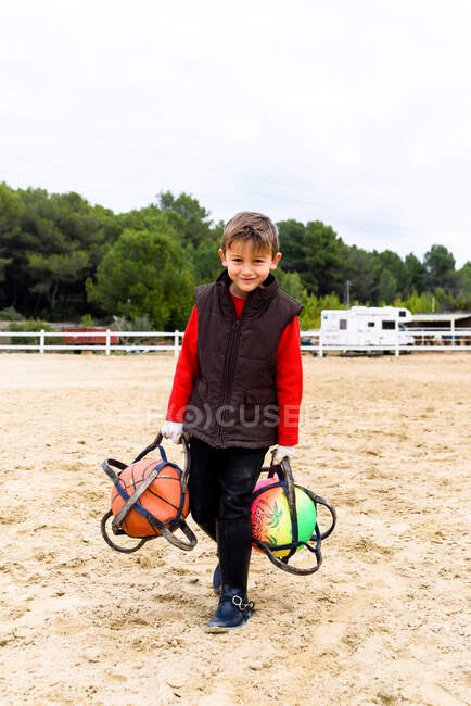 Corpo inteiro alegre pequeno jóquei carregando bolas de treinamento e olhando para a câmera enquanto caminhava na arena de curativo na escola equestre — Fotografia de Stock
