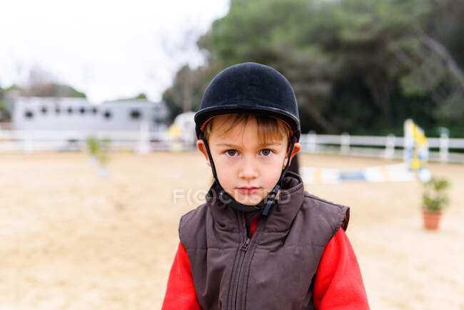Netter kleiner Jockey in Helm und Weste, der in die Kamera schaut, während er auf dem verschwommenen Hintergrund der Pferdekoppel der Reitschule steht — Stockfoto