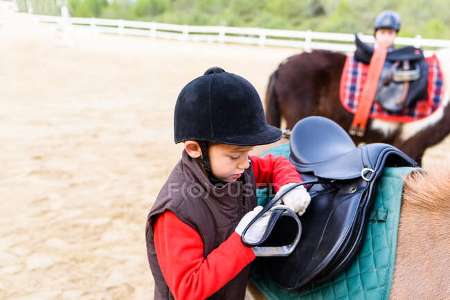 Vue latérale du petit jockey en casque de protection ajustant l'étrier sur la selle avant de monter poney asymétrique à l'école équestre — Photo de stock