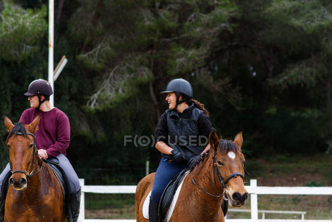 Счастливые подростки-жокеи в шлемах общаются друг с другом, катаясь на покорных лошадях на песчаной арене во время урока в конной школе — стоковое фото