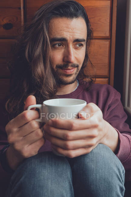 Бородатий дорослий чоловік з довгим волоссям насолоджується гарячим напоєм з білого кубку і дивиться в сторону, відпочиваючи в затишній кімнаті вдома — стокове фото