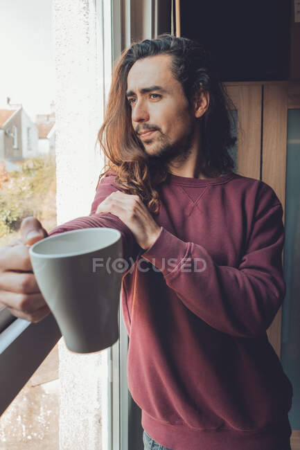 Hombre barbudo adulto con cabello largo disfrutando de una bebida caliente y mirando por la ventana mientras pasa tiempo en casa - foto de stock
