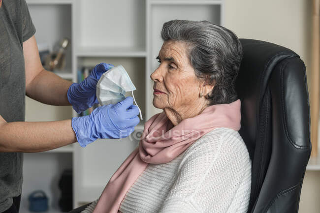 Социальный работник в латексных перчатках надевает медицинскую маску на пожилую женщину на стул, заботясь о пенсионере во время пандемии коронавируса дома — стоковое фото