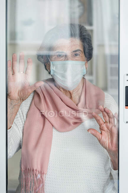 Anciana con máscara médica mirando a la cámara a través de las ventanas mientras permanece en casa durante la epidemia de coronavirus - foto de stock