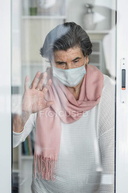 Anciana con máscara médica mirando a través de las ventanas mientras permanece en casa durante la epidemia de coronavirus - foto de stock