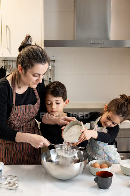 Братья и сёстры с миской муки улыбаются, помогая матери в фартуке готовить тесто на уютной кухне дома — стоковое фото