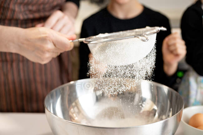 Неузнаваемая женщина просеивает муку в металлическую миску во время приготовления выпечки с детьми на кухне дома — стоковое фото