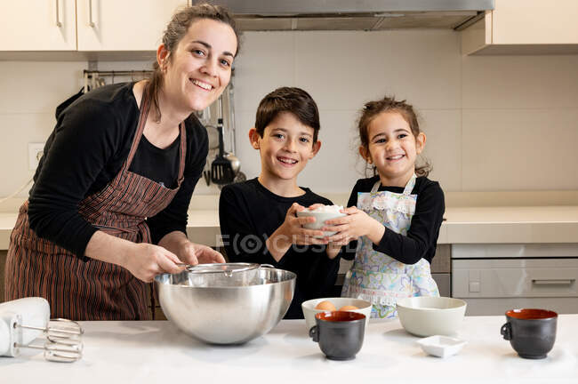 Geschwister mit einer Schüssel Mehl lächeln, während sie der Mutter in der Schürze helfen, in der gemütlichen Küche zu Hause Gebäck zuzubereiten — Stockfoto
