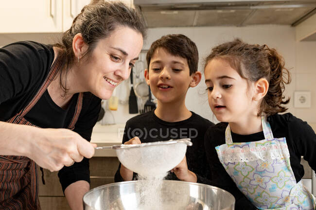 Los hermanos con tazón de harina sonriendo mientras que ayuda a la madre en el delantal para preparar pastelería en la acogedora cocina en casa - foto de stock
