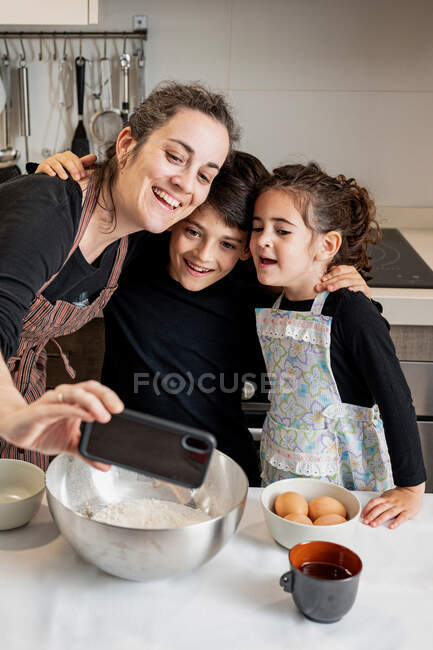 Donna felice in grembiule sorridente e scattare selfie con il telefono cellulare con bambini felici durante la cottura pasticceria insieme in cucina accogliente a casa — Foto stock