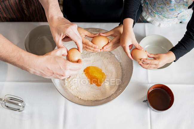 From above anonymous parent and little siblings cracking and adding raw eggs to flour in metal bowl while preparing pastry dough in kitchen together — Stock Photo
