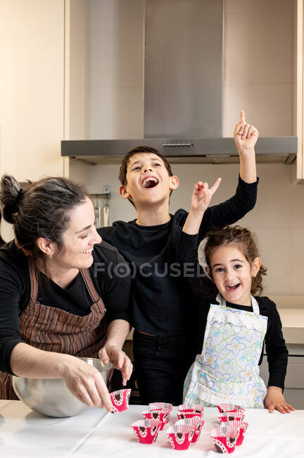 Happy adult woman smiling and looking at excited children pointing up while cooking cupcakes at home together — Stock Photo