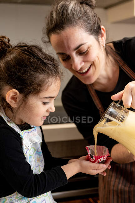 Mujer feliz sonriendo y vertiendo masa en la taza de papel mientras prepara cupcakes con su hija pequeña en el día de fin de semana en casa - foto de stock