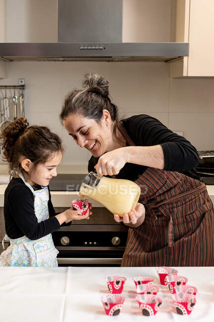 Femme heureuse souriant et verser de la pâte dans une tasse en papier tout en préparant des cupcakes avec petite fille le week-end à la maison — Photo de stock