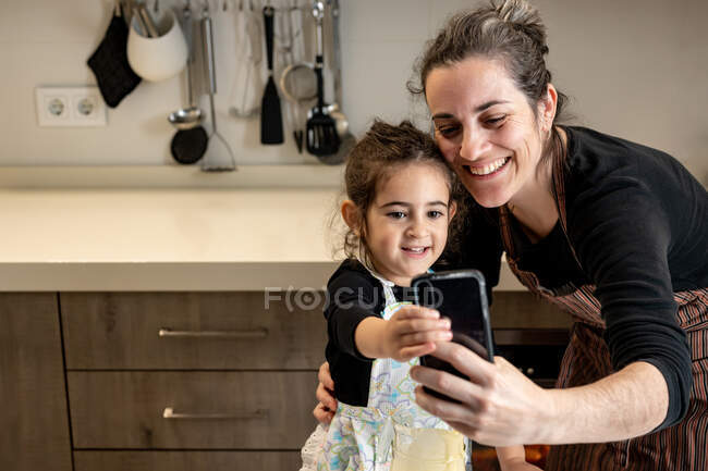 Счастливая женщина в фартуке улыбается и делает селфи с мобильным телефоном со счастливой маленькой девочкой, готовя вместе кондитерские изделия на уютной кухне дома — стоковое фото