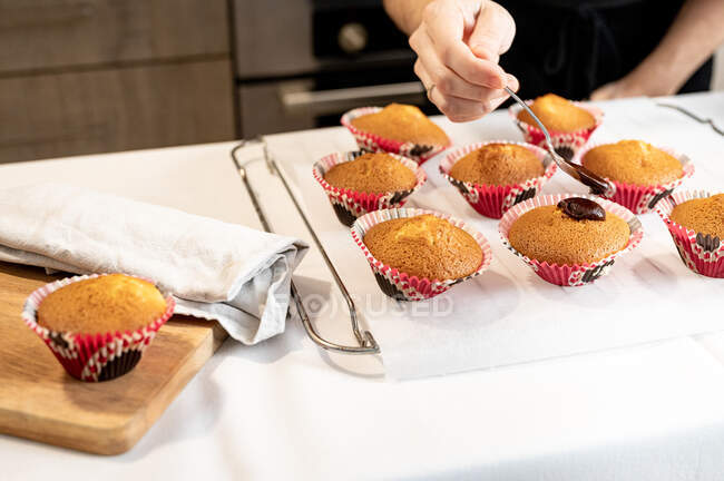 Mujer de la cosecha usando cuchara para esparcir glaseado de chocolate en cupcakes recién horneados mientras prepara pasteles en casa - foto de stock