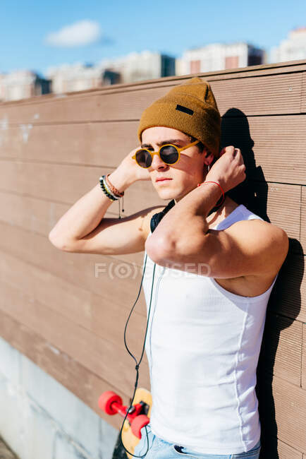 Young handsome male hipster with headphones leaning on wall near skateboard before listening to music on sunny day in city — Stock Photo