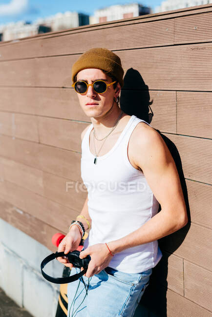 Joven guapo hipster masculino con auriculares apoyados en la pared cerca del monopatín antes de escuchar música en un día soleado en la ciudad - foto de stock