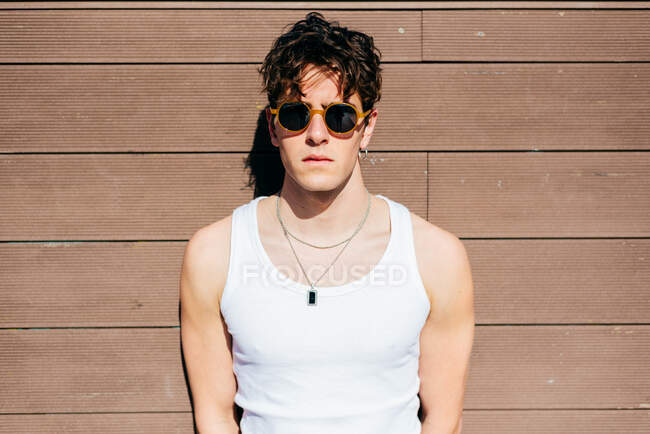 Сучасний молодий красивий чоловік в модних сонцезахисних окулярах і білий танк зверху стоїть біля коричневої стіни в сонячний день на вулиці міста — стокове фото