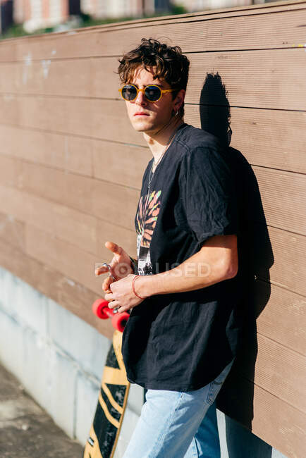 Красивий чоловічий хіпстер в сонцезахисних окулярах, що стоїть біля скейтборду в сонячний день на вулиці міста — стокове фото