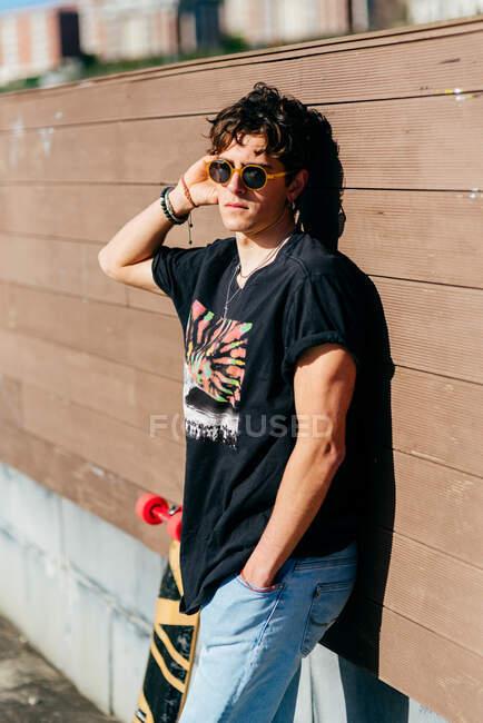 Красивый хипстер в солнечных очках, стоящий возле скейтборда в солнечный день на городской улице — стоковое фото