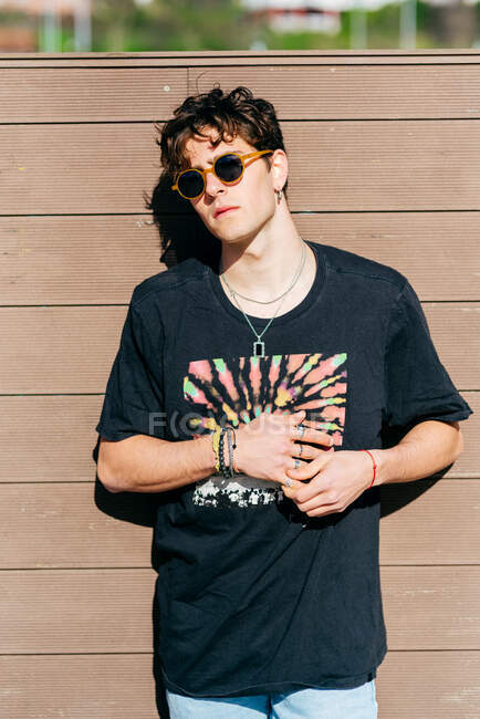 Joven hombre guapo moderno en gafas de sol de moda y camiseta negra de pie cerca de la pared marrón en el día soleado en la calle de la ciudad - foto de stock