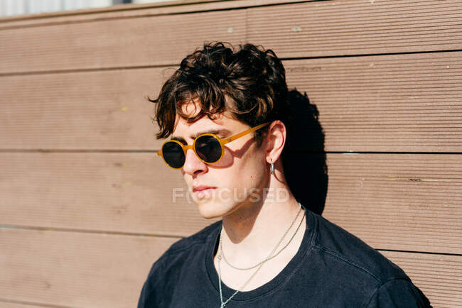 Jeune homme moderne beau dans des lunettes de soleil à la mode et t-shirt noir debout près du mur brun par une journée ensoleillée sur la rue de la ville — Photo de stock