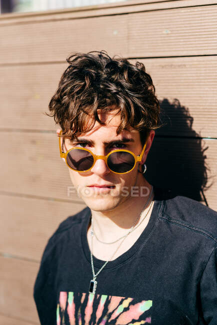 Joven hombre guapo moderno en gafas de sol de moda y camiseta negra de pie cerca de la pared marrón en el día soleado en la calle de la ciudad - foto de stock