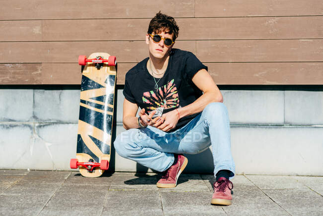 Handsome male hipster in sunglasses sitting on haunches near skateboard on sunny day on city street — Stock Photo