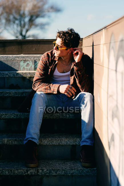 Молодой красивый парень в повседневной одежде трогает ухо и смотрит в сторону, сидя на лестнице в солнечный день на городской улице — стоковое фото