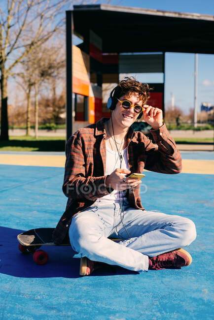 Happy hipster in casual clothes smiling for camera and browsing smartphone while sitting on skateboard and listening to music on sports ground — Stock Photo