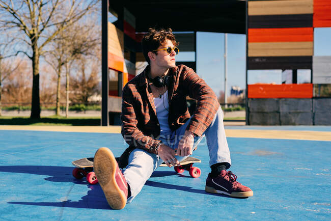 Confident young handsome man in casual clothes sitting on skateboard and looking away on sunny day in skate park — Stock Photo