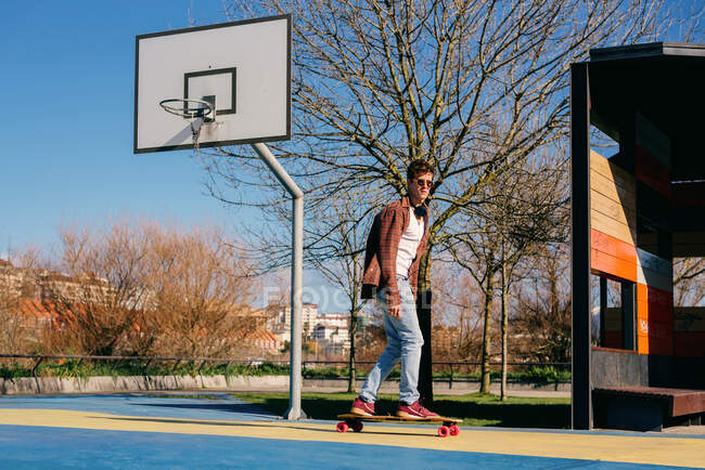 Full body young man in casual clothes riding skateboard near basketball hoop on sunny day on sports ground in city — Stock Photo