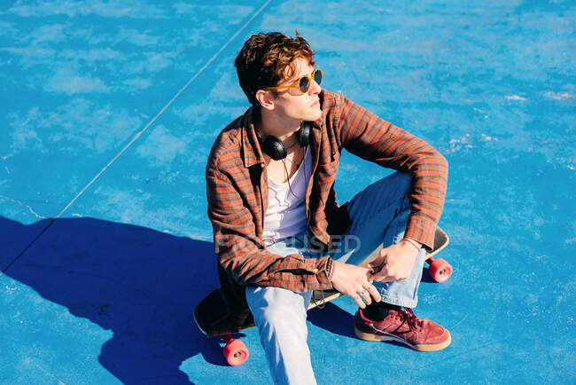 From above confident young man in casual clothes sitting on skateboard and looking away on sunny day in skate park — Stock Photo