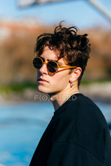 Beau jeune homme en t-shirt foncé, lunettes de soleil et boucles d'oreilles regardant la caméra au-dessus de l'épaule tout en se tenant debout sur fond flou de la rue de la ville — Photo de stock