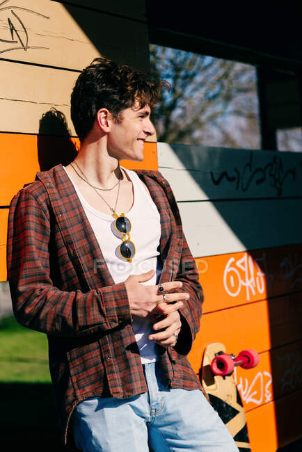 Handsome male hipster standing near skateboard looking away on sunny day on leaning on a wooden colorful wall in the city street — Stock Photo