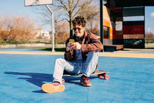 Счастливый хипстер в повседневной одежде улыбается и просматривает смартфон, сидя на скейтборде и слушая музыку на спортивной площадке — стоковое фото
