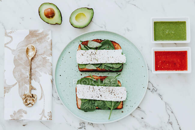 From above plate with cheese and spinach sandwiches place on marble tabletop near napkin with spoon and bowls with avocado and tomato sauces — Stock Photo