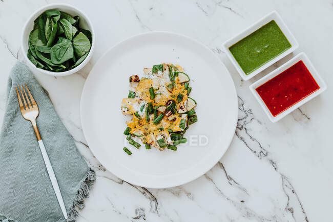 From above plate with zucchini and green beans salad with cheese placed on marble table near bowl with fresh spinach leaves and avocado and tomato sauces next to napkin with fork — Stock Photo