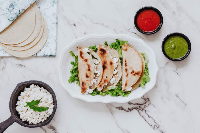 Tortillas mit frischem Quark auf Teller mit Salatblättern in der Nähe von Schüsseln mit Avocado und Tomatensaucen auf Marmortisch — Stockfoto