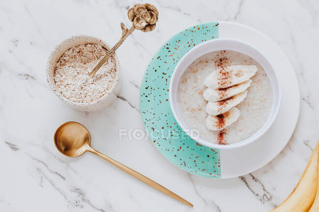 Top view of bowl of healthy oatmeal porridge served with slices of banana and cinnamon near spoon on marble tabletop — Stock Photo