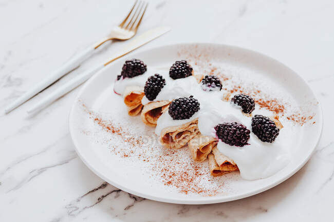 Delicious crepes with yoghurt and blackberries served on plate with cinnamon near silverware on marble table — Stock Photo
