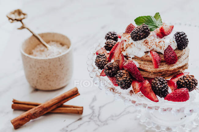 Deliciosos panqueques con fresas frescas y moras decoradas con hojas de menta colocadas en el plato cerca de palitos de canela y una taza de helado en la mesa de mármol - foto de stock