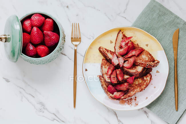 Top view of delicious egg fried bread with ripe strawberries served on plate near cutlery and napkin on marble tabletop — Stock Photo