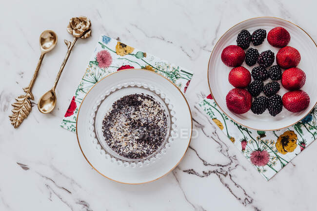 Bowl with delicious poppy seed pudding and plate with strawberries and blackberries placed near paper napkins and ornamental spoons on marble table — Stock Photo