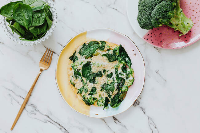 Von oben leckerer Brokkoli-Salat mit Spinatblättern und geriebenem Käse, serviert auf einem Teller in der Nähe einer Gabel auf einer Marmorplatte — Stockfoto