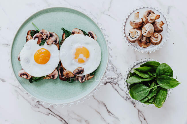 Top view of tasty fried eggs placed of yummy sandwiches with mushrooms and spinach on plate on marble table — Stock Photo