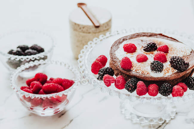 Высокий угол вкусного сладкого пирога со сливочной начинкой и свежей ежевикой и малиной на стеклянном стенде в составе с чашами свежих ягод — стоковое фото