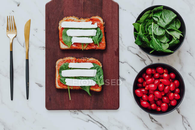Vista superior de torradas de pão com espinafre verde e queijo mussarela em molho de tomate composto em placa de madeira com tigelas de ingredientes e talheres na superfície de mármore — Fotografia de Stock